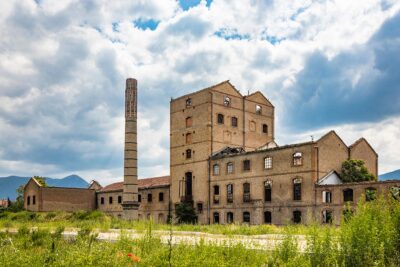 An old disused factory, abandoned and in ruins, with a smashed roof and a chimney. Tall weeds invade the building. Concept of economic bankruptcy. Cloudy sky. Italy, Foligno, Umbria.
